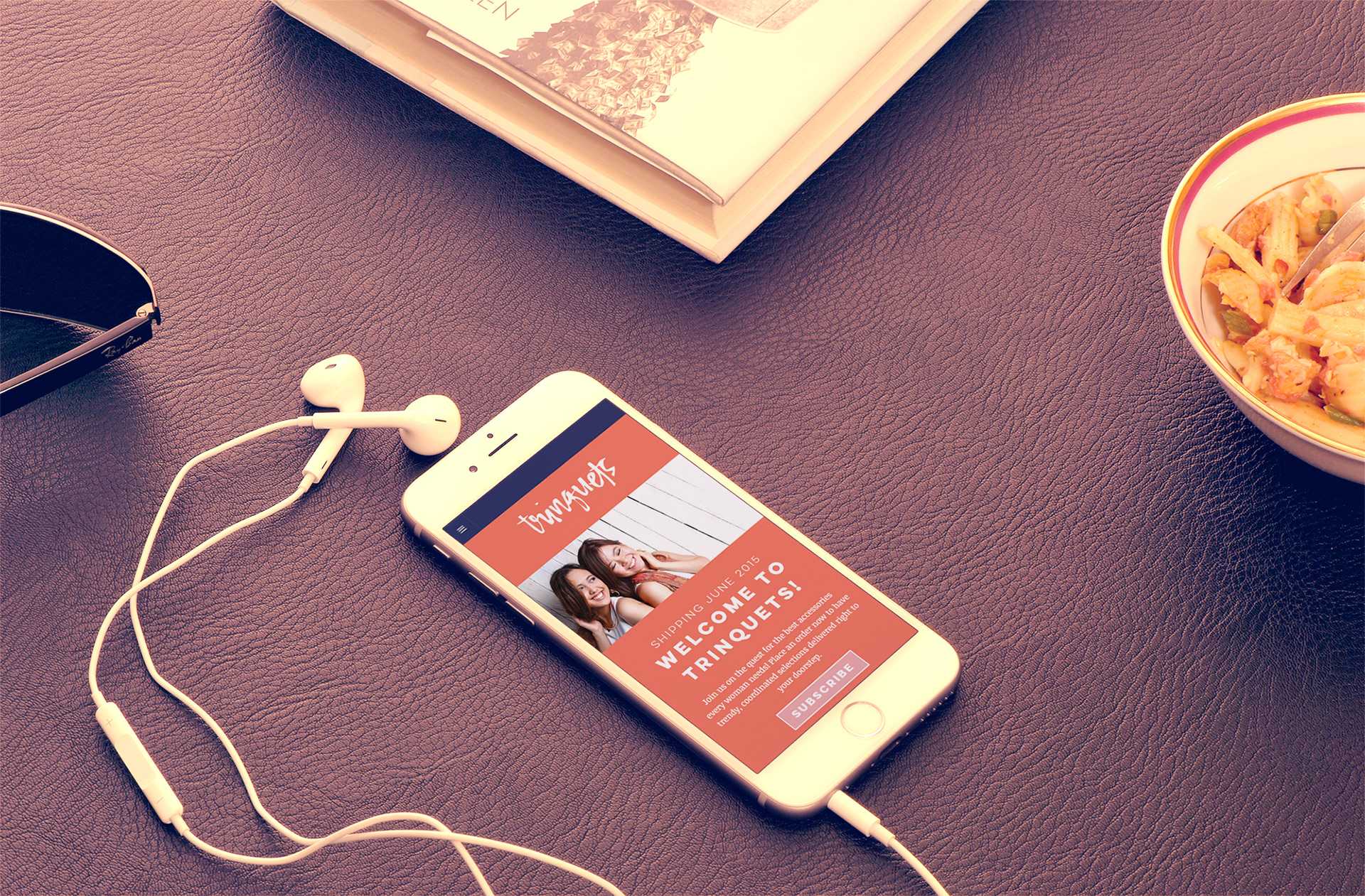 The Trinquets mobile site shown on a smartphone in a flat-lay photo with earphones, a magazine, sunglasses, and a bowl of pasta